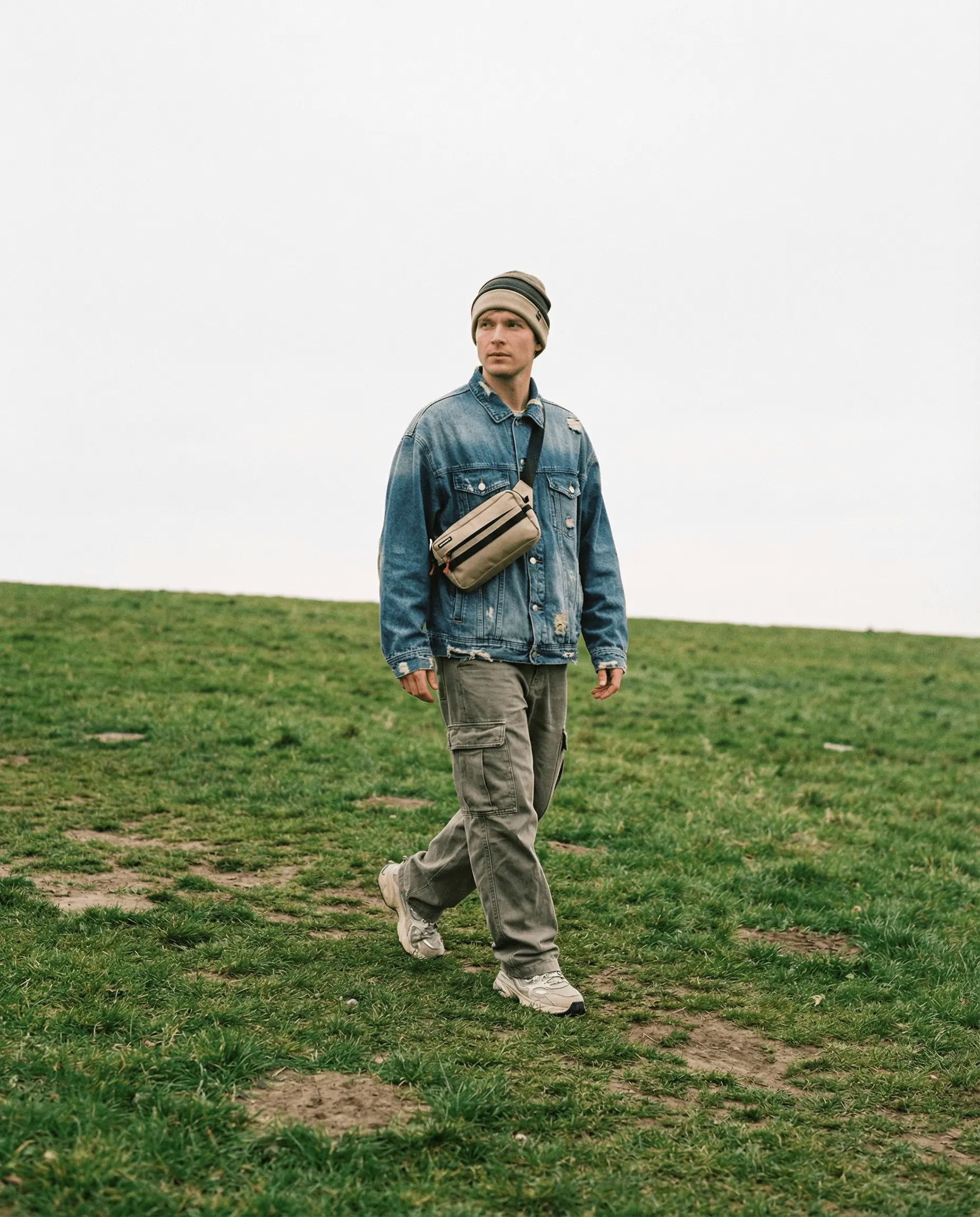 Muž v modré džínové bundě a čepici, nosí crossbody ledvinka cordura na zelené trávě.